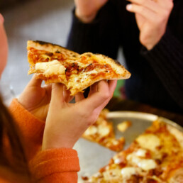 Close-up of a female customer holding a slice of pizza in her hands, with another customer sitting across from her