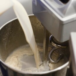 Yeast and water mixture being poured from a plastic jug into the bowl of a large metal dough-mixer