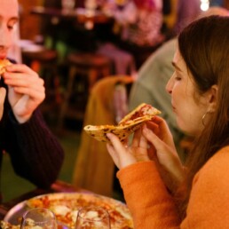 Male customer and female customer sitting opposite eachother, eating pizza slices they are holding in their hands