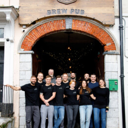 Close-up of Pompeii Pizza staff & Franciscan Well Brewery & Brew Pub staff members, standing in front of the Franciscan Well entrance arch