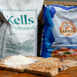 Mound of grain and mound of milled flour on wooden plates with a bag of Kells Wholemeal grain and a bag of Caputo flour in the background